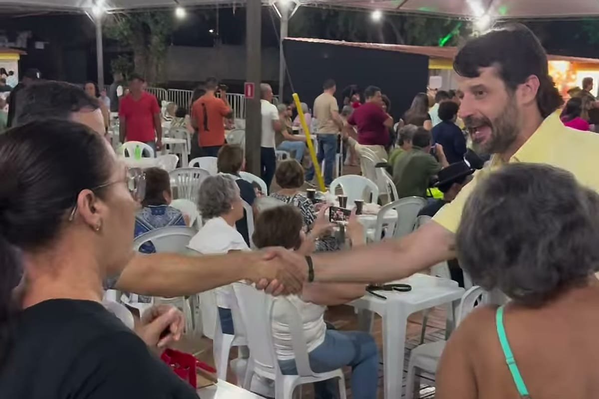 Guilherme Piai, de camisa amarela, aperta a mão de uma mulher em um evento noturno, com mesas e pessoas sentadas ao fundo