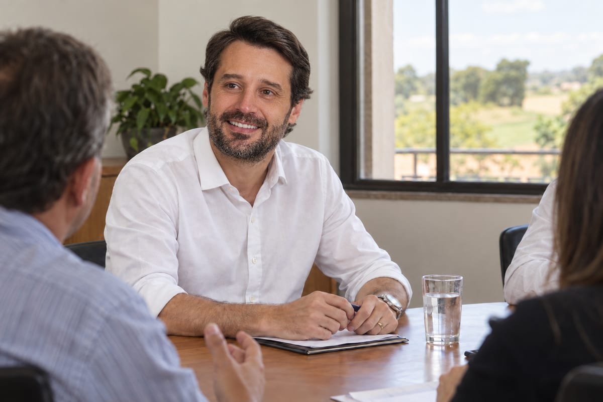 Guilherme Piai, de camisa social branca, sentado à mesa de uma sala de reunião, com uma pasta e uma caneta, ouvindo duas pessoas de costas, ao lado de uma janela
