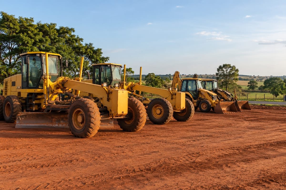 Motoniveladoras e retroescavadeiras amarelas estacionadas em terra vermelha, com árvores e pasto ao fundo