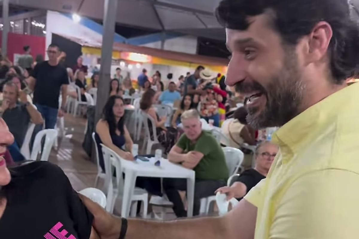 Guilherme Piai, de camisa amarela, cumprimenta uma mulher em um evento noturno, com mesas e pessoas sentadas ao fundo