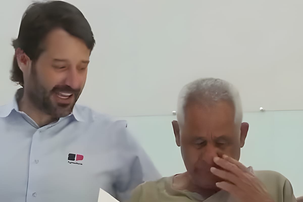 Guilherme Piai, de camisa clara, sorri olhando para um papel ao lado de um senhor idoso de cabelos grisalhos que leva a mão ao rosto