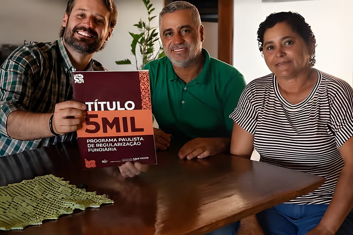 Guilherme Piai, de camisa xadrez, segura uma publicação com os dizeres "Título 5 mil, Programa Paulista de Regularização Fundiária", ao lado de um homem de camisa verde e de uma mulher de blusa listrada, sentados a uma mesa